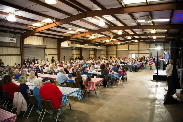 A packed hall of guests at checked tablecloths, with a speaker at the podium