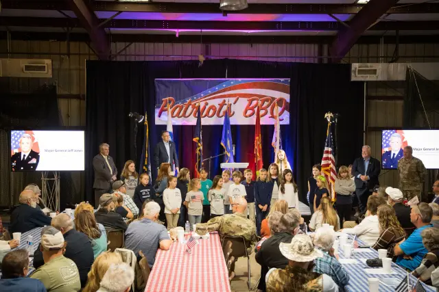 Children stand on stage under the Patriots' BBQ banner while guests watch