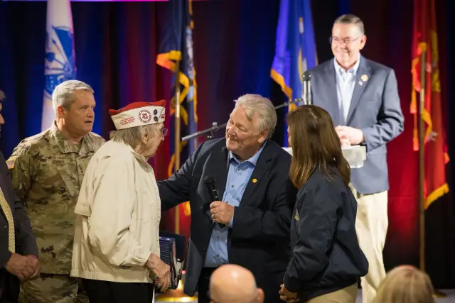 A veteran in a decorated cap is greeted on stage